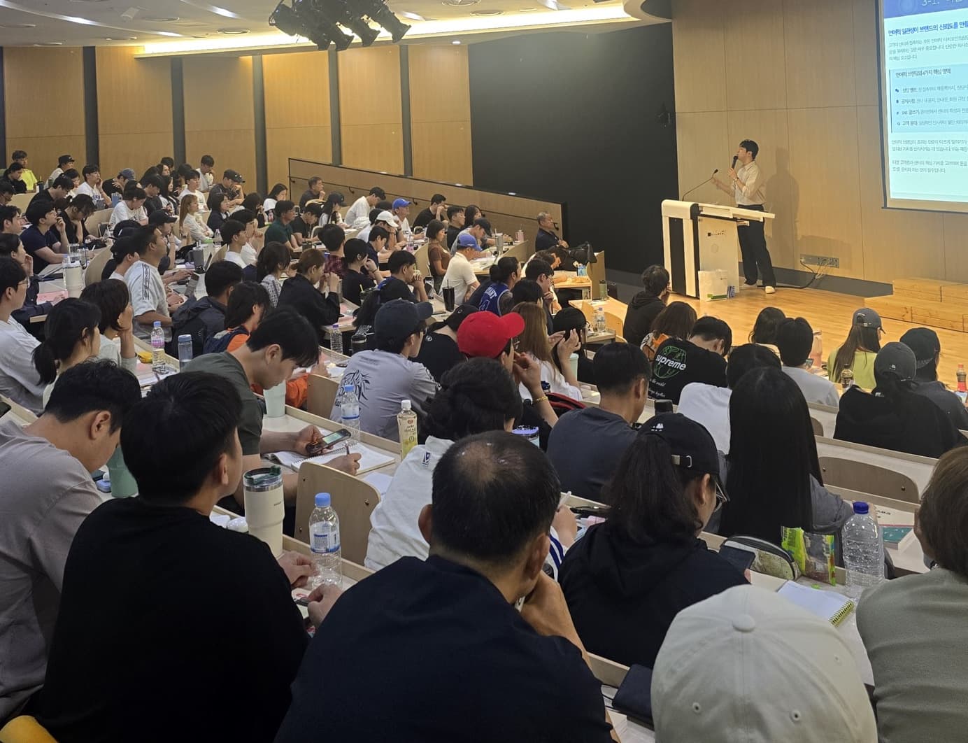 강당에서 강연자가 발표 화면 옆에 서 있고 수강생들이 자리를 채워 앉은 장면